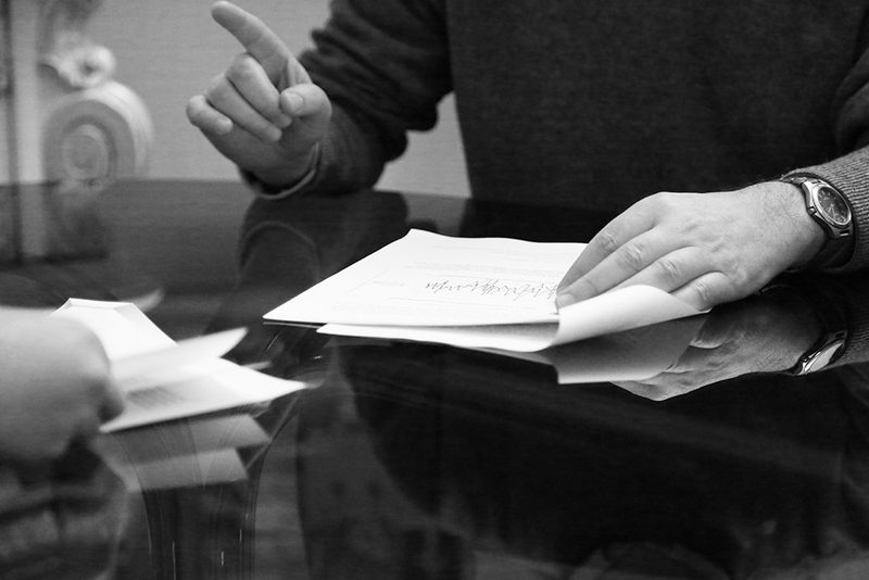 Svartvitt foto: en man håller en bunt papper med ett linjediagram mot ett blankt mötesbord och pekar med fingret medan en annan persons hand räcker fram egna papper från andra sidan bordet.